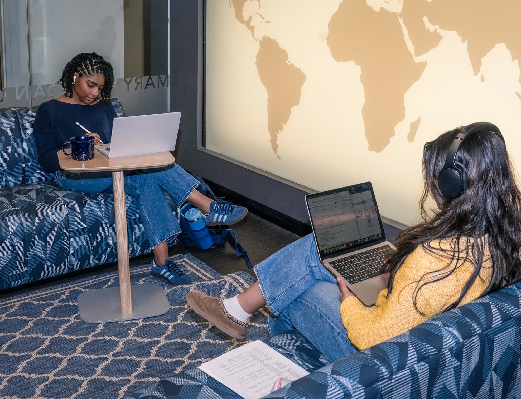 Two students studying on laptops in a lounge near a world map mural